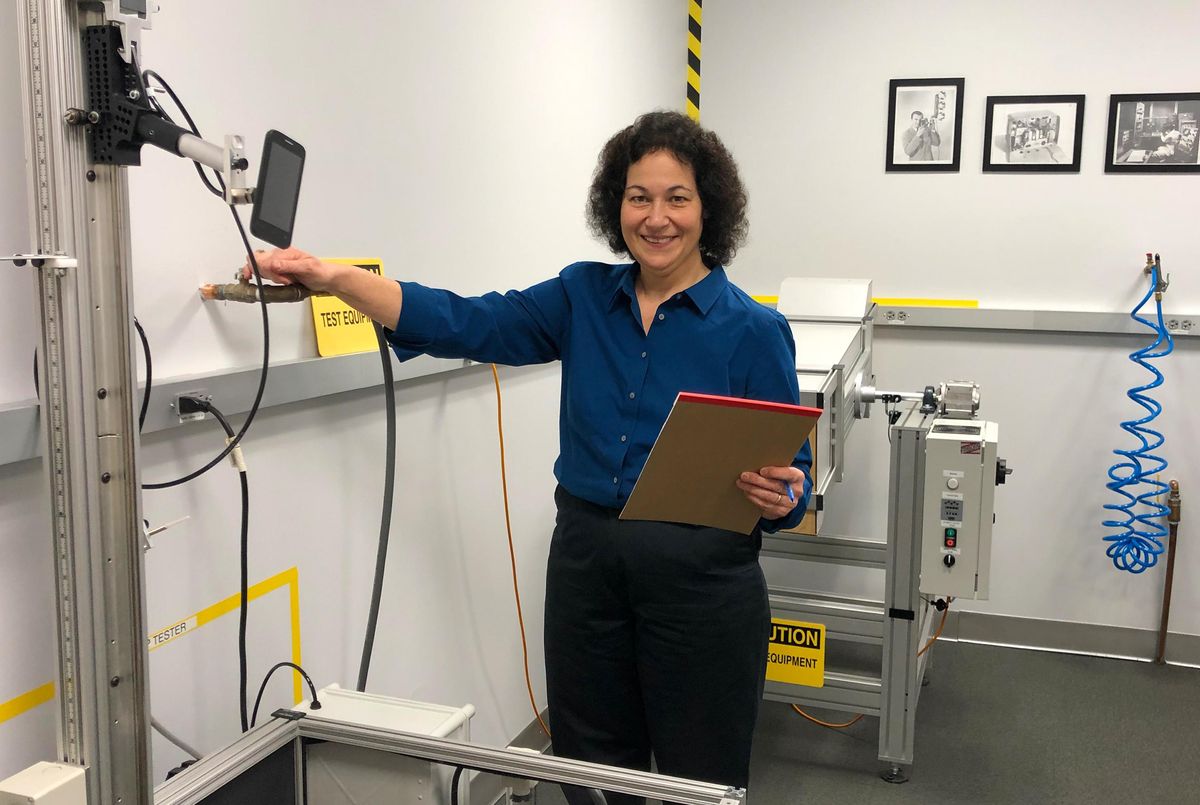Maria Rerecich在《消费者报告》位于纽约州扬克斯的实验室里，带着手机掉落塔(前面)和玻璃杯(后面)测试设备。