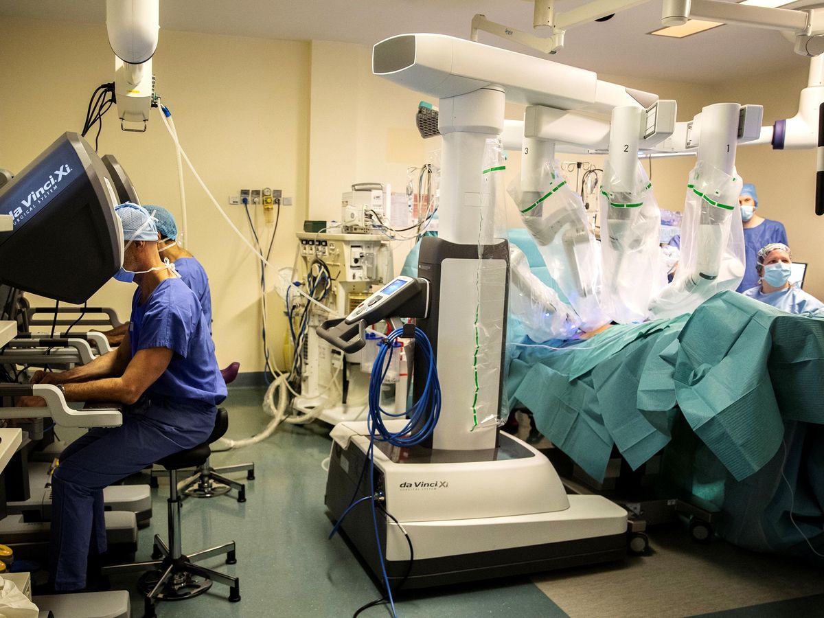手术室照片。在图像的左侧，两名外科医生坐在控制台前，双手放在控制台上。在右侧，一个有四只手臂的白色大型机器人正在给病人做手术。