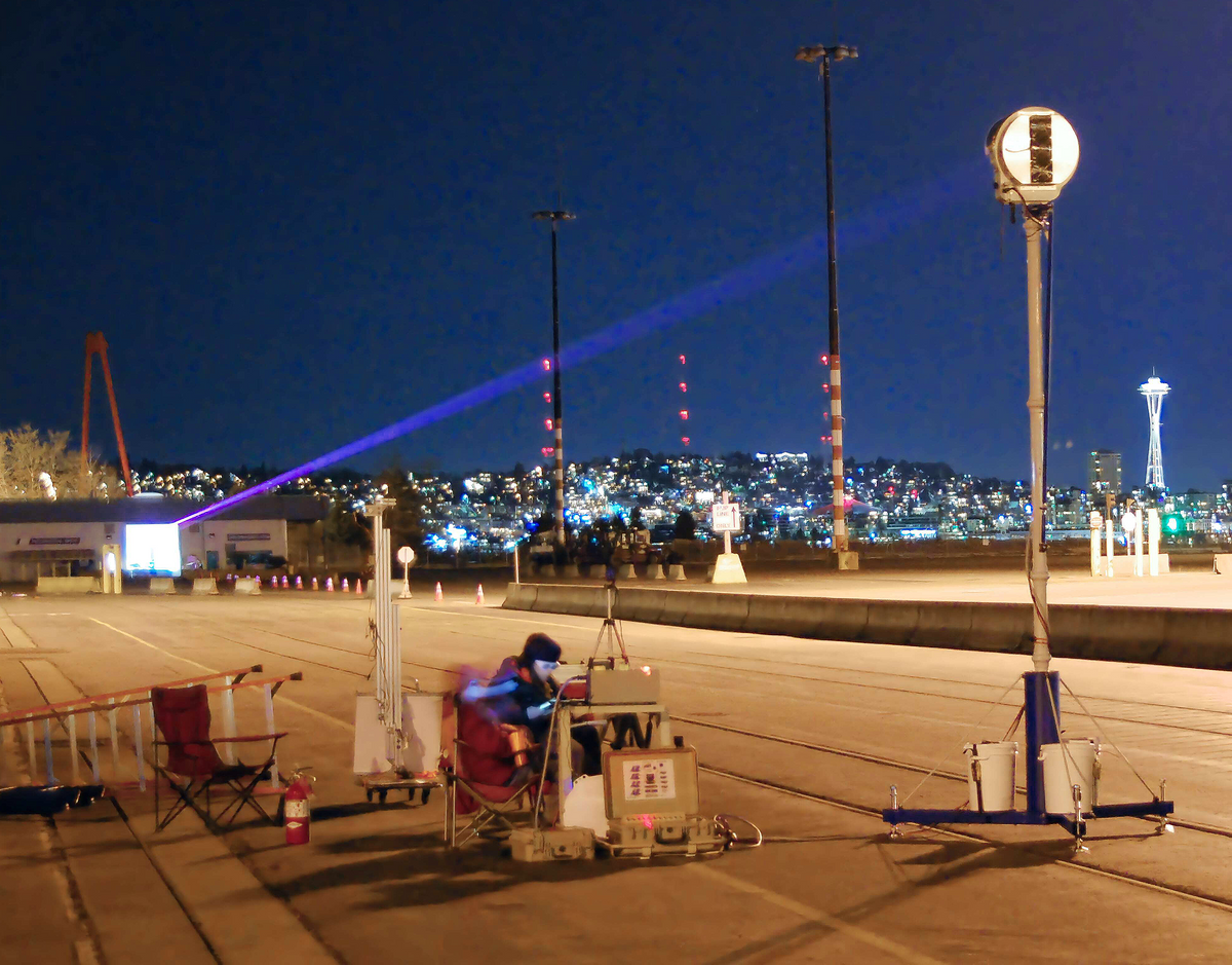 这张夜间户外照片，背景是城市的灯光，显示出一束狭窄的光束照射在一个位于杆子顶部的圆形接收器上。