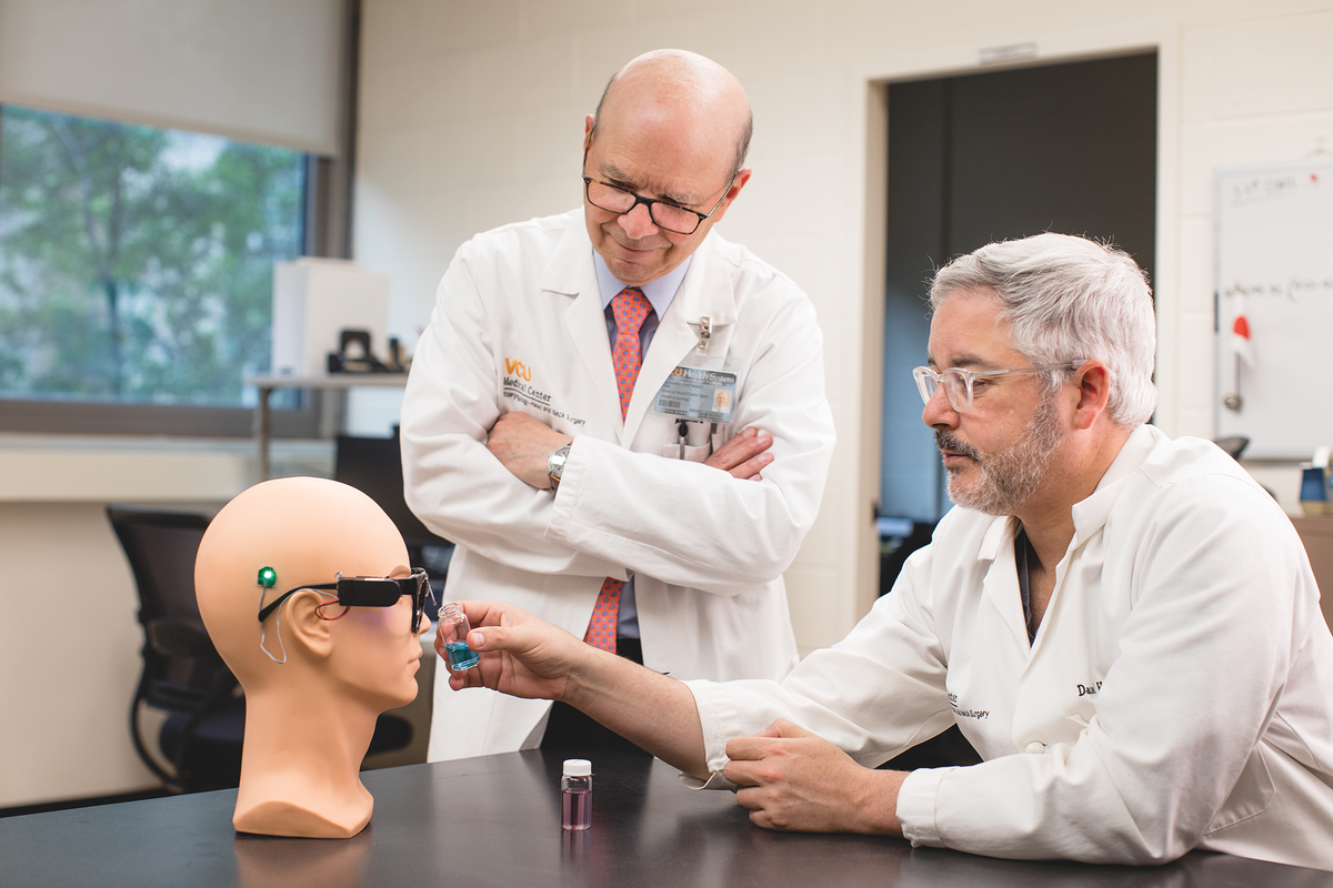 两名身穿白大褂的科学家正在观察一个戴着一副黑色眼镜、几处有电线伸出的人体模型头部。其中一名科学家将一小瓶蓝色液体举到鼻梁上方的电线上。桌上放着一瓶紫色的液体。