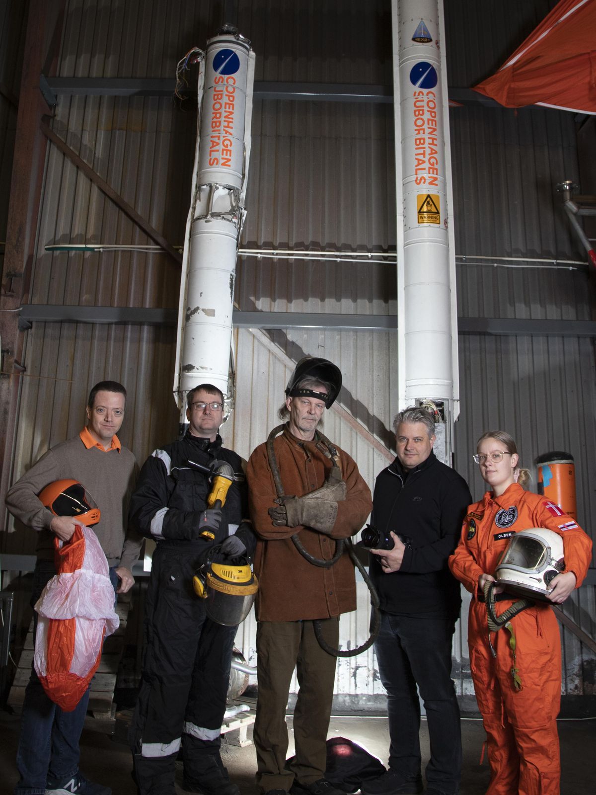 五个人站在两个高大的火箭前面。一些人穿着宇航服，拿着头盔，另一些人拿着焊接设备。＂aria-label=